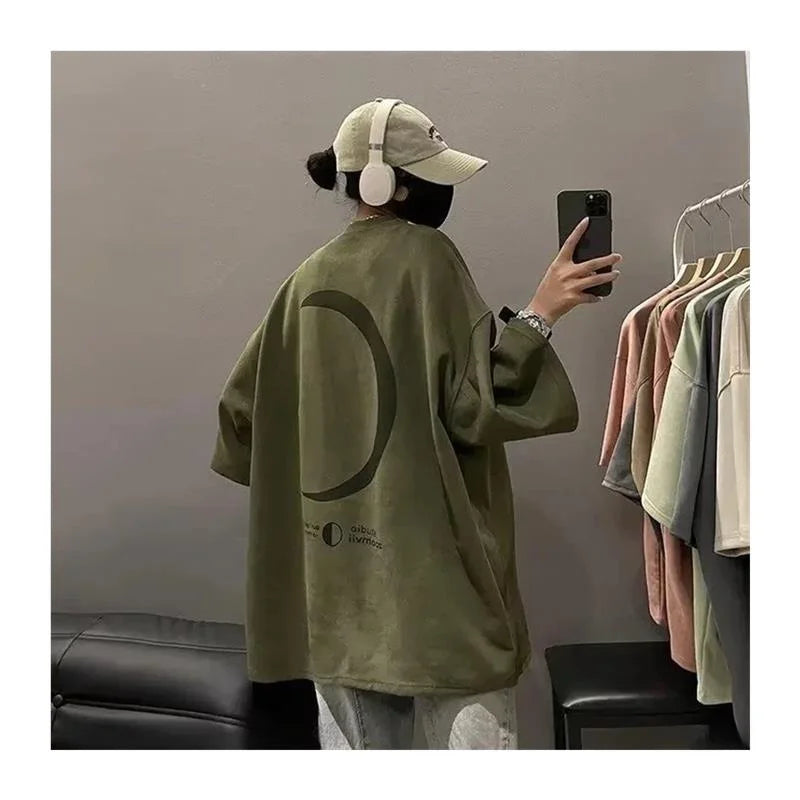 Person wearing a green oversized shirt with a logo, taking a mirror selfie in a room with clothing racks.