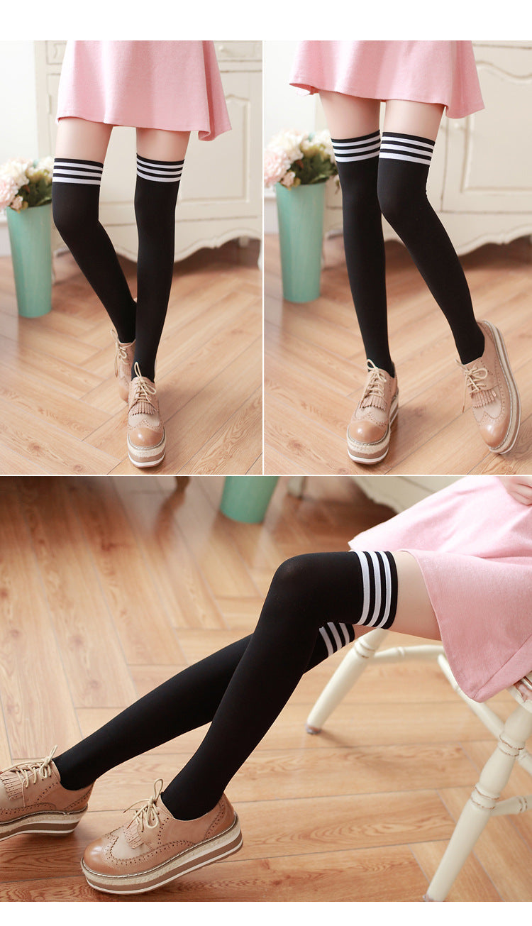 Black knee-high socks with white stripes worn by a person in a pink skirt and beige shoes on a wooden floor.