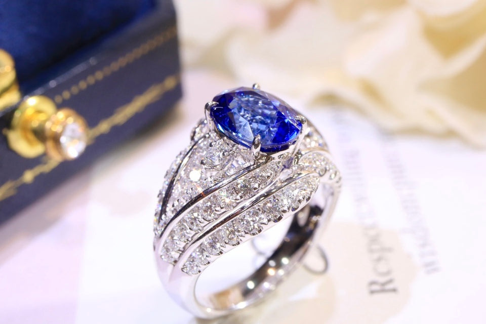 A silver-toned ring featuring a large oval-cut deep blue gemstone, set with multiple rows of small clear stones on a white surface, with a dark blue jewelry box and soft cream flowers in the background.