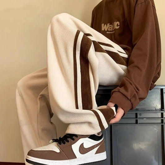 Person wearing a brown sweatshirt and beige pants with white stripes, standing against a neutral background.