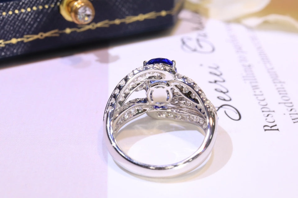 A silver-toned ring with a central deep blue oval gemstone and smaller clear stones, photographed on a white surface with a blurred dark blue fabric and printed card in the background.