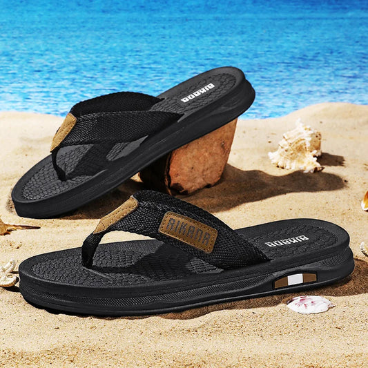 Black sandals with brown accents on a sandy beach with blue water in the background