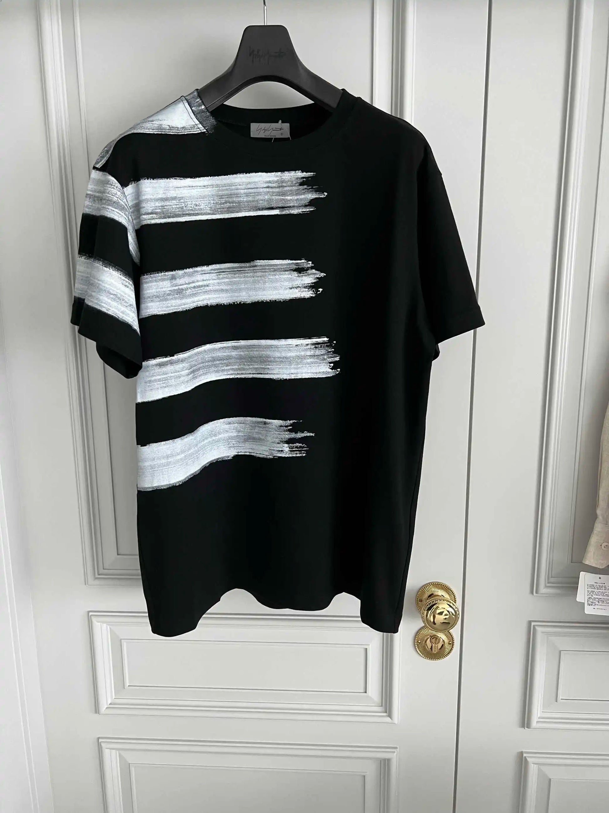 Black t-shirt with white brushstroke design on a hanger against a white door.