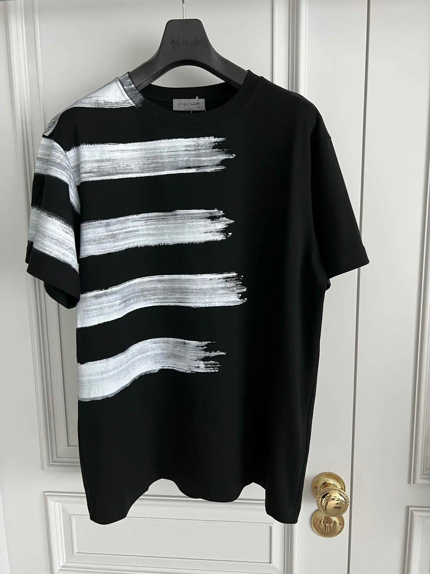 Black t-shirt with white brushstroke design on a hanger against a white door.
