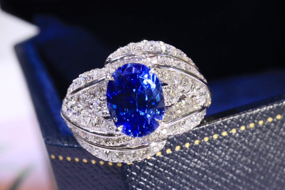 An oval-cut blue sapphire ring set in a silver-colored metal with diamond pavé bands, displayed on a dark textured jewelry box.