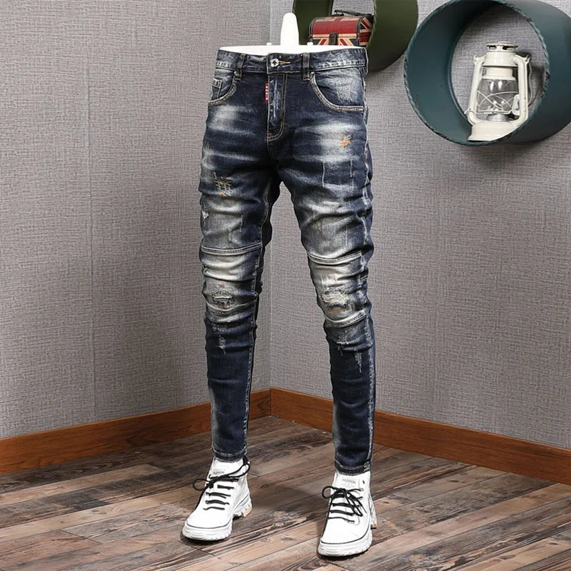 Distressed blue jeans worn by a person on a wooden floor with a gray textured wall.