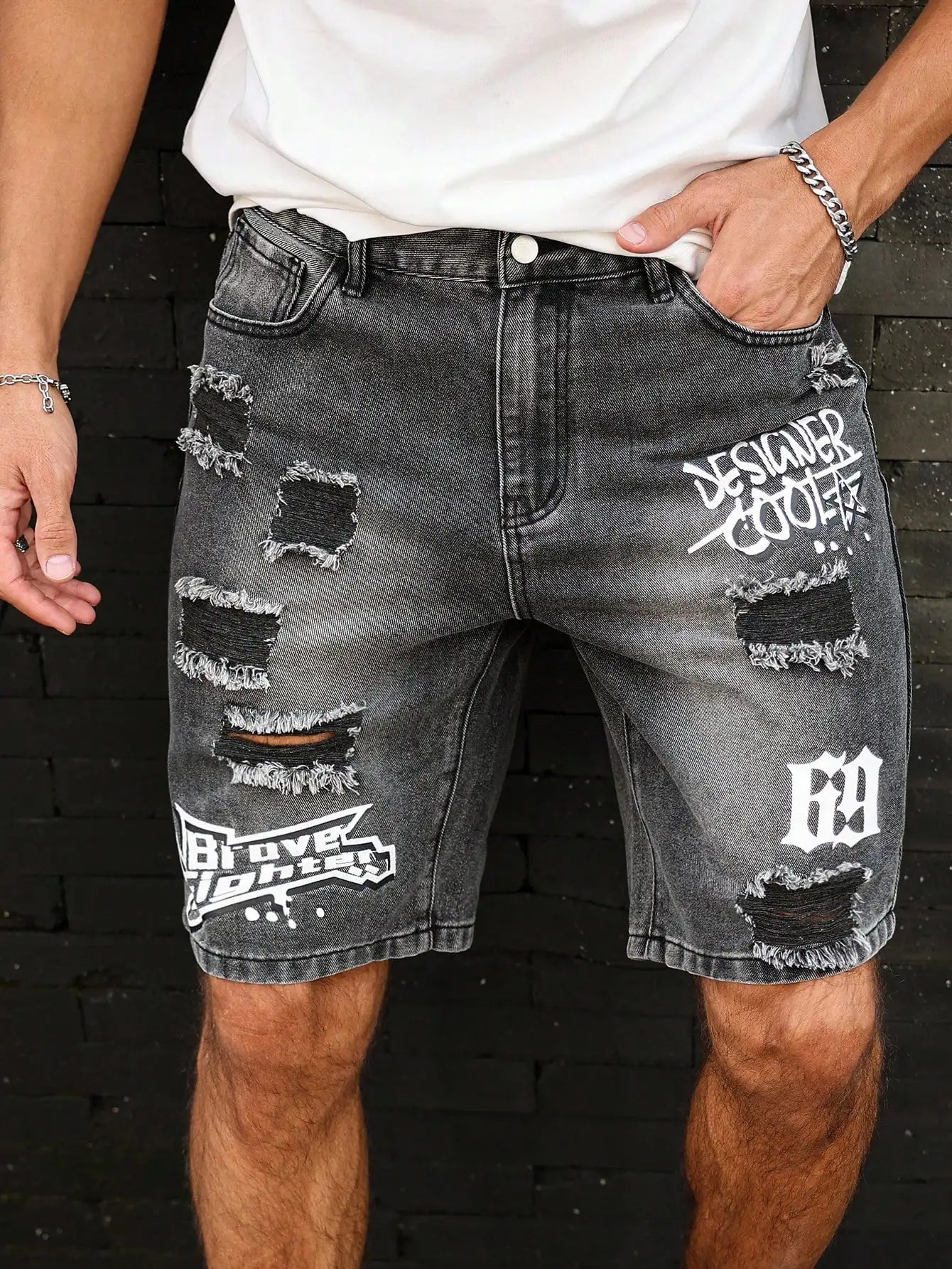 Person wearing black distressed denim shorts with branding against a dark brick wall.