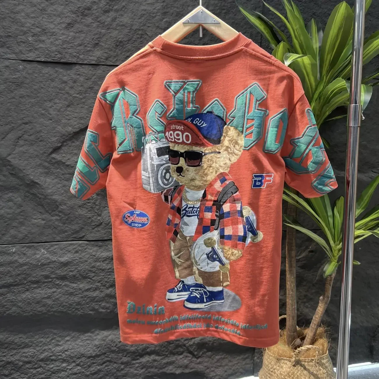 Orange t-shirt with graphic design of a bear wearing sunglasses and a cap, hanging against a dark textured wall.