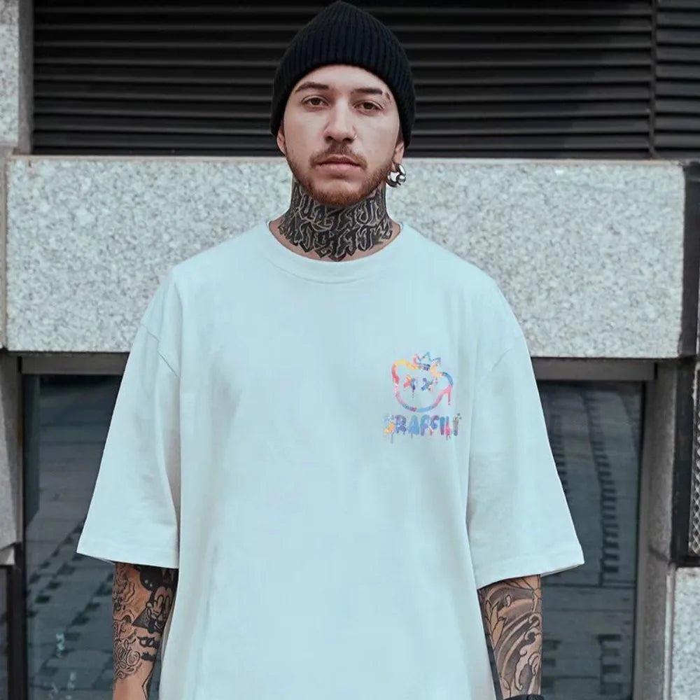 Man wearing a light blue t-shirt with a colorful logo and text, standing against a textured wall.