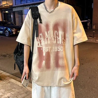 Person wearing a beige t-shirt with text, holding a phone, and standing on a street.
