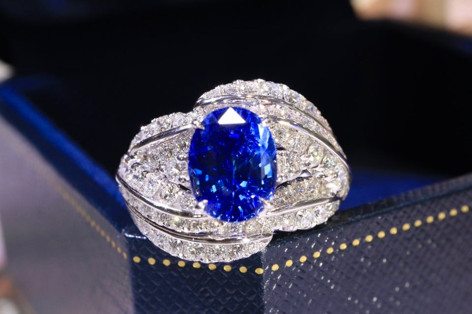 A silver-toned ring featuring a large oval-cut vivid blue gemstone, surrounded by pavé-set small clear stones, displayed on a dark blue jewelry box.