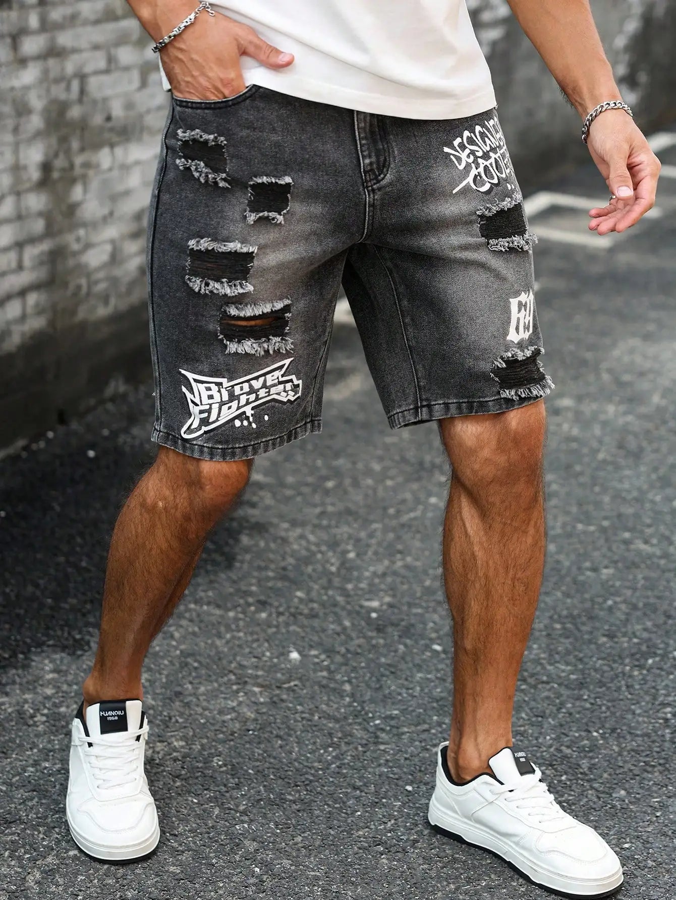 Person wearing black distressed denim shorts with white text and white sneakers on a street.