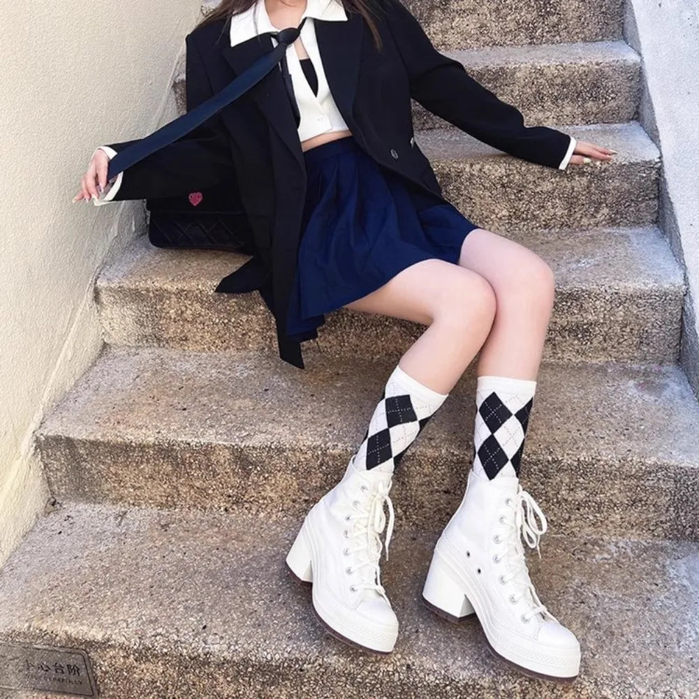 Person sitting on stairs wearing a black coat, white shirt, blue skirt, and white boots with black diamond patterns.