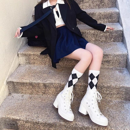 Person sitting on stairs wearing a black coat, white shirt, blue skirt, and white boots with black diamond patterns.