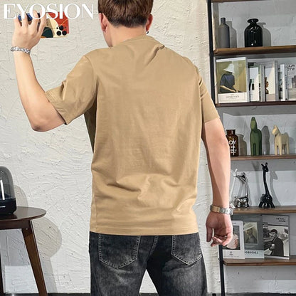 Man wearing a beige t-shirt taking a selfie in a room with shelves and decor.