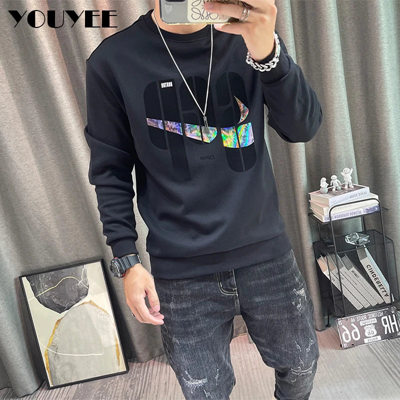 Person wearing a black sweatshirt with colorful design and jeans, standing in front of a curtain.