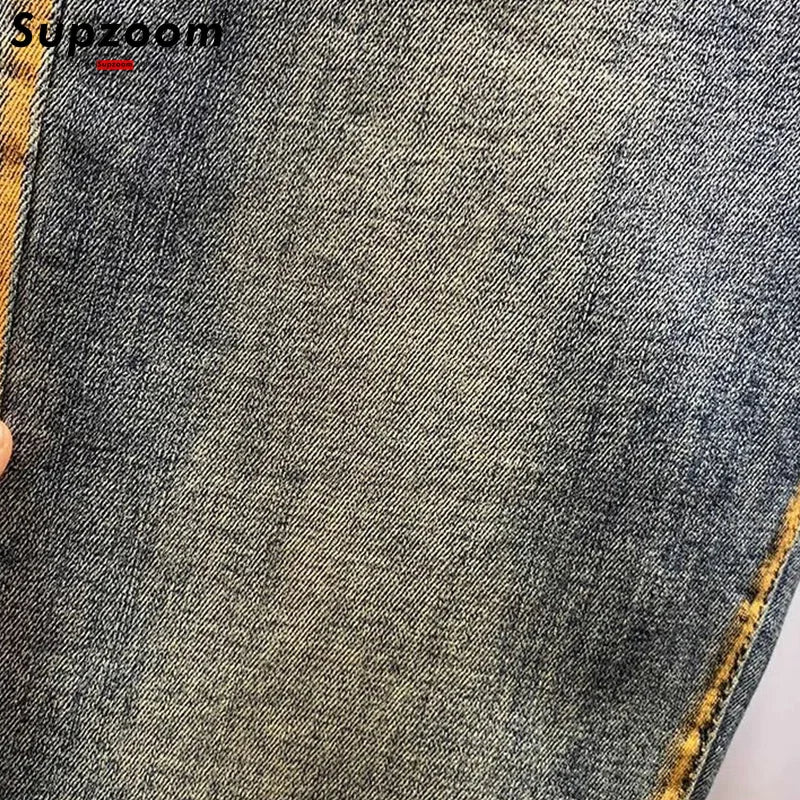 Close-up of gray denim fabric with visible stitching details, brand 'Supzoom' in top left corner.