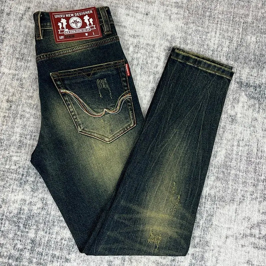 A pair of folded dark blue denim jeans with a faded yellow-green wash, laid flat on a textured gray background. The back pocket of the left leg shows a curved double-stitch detail, and the red 'DICKI NEW DESIGNER' brand patch is visible at the waistband.