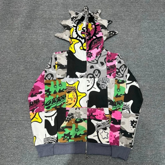 A full-zip hoodie featuring a vibrant graffiti patchwork design with multiple colors including pink, green, yellow, black, and white, laid flat on a gray textured background.