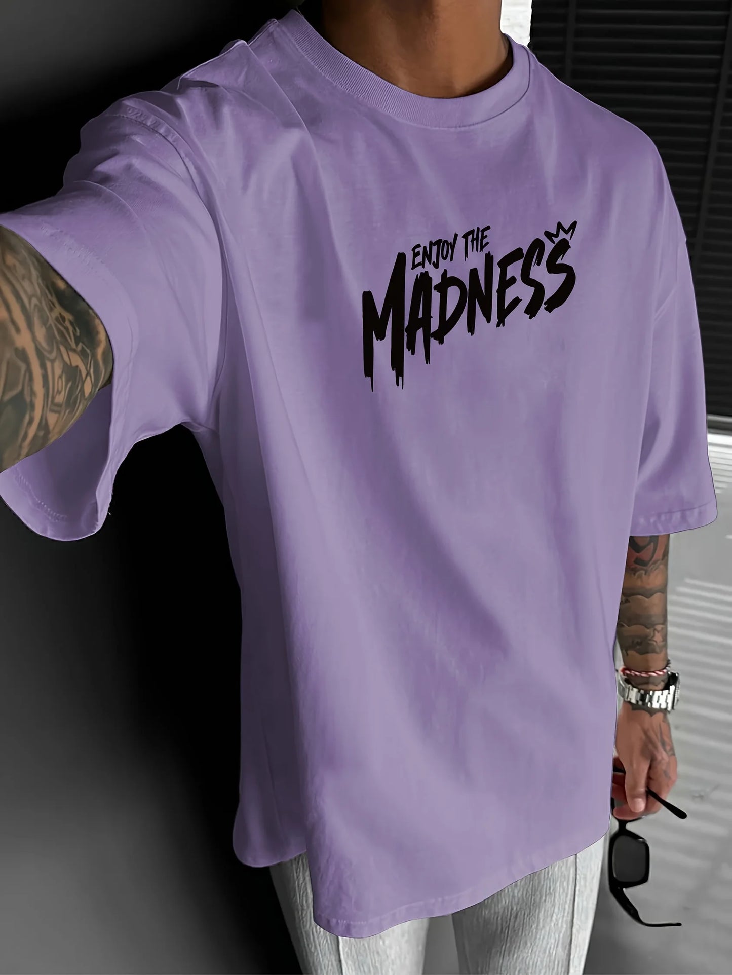 Person wearing a purple t-shirt with 'Enjoy the Madness' text, holding sunglasses.
