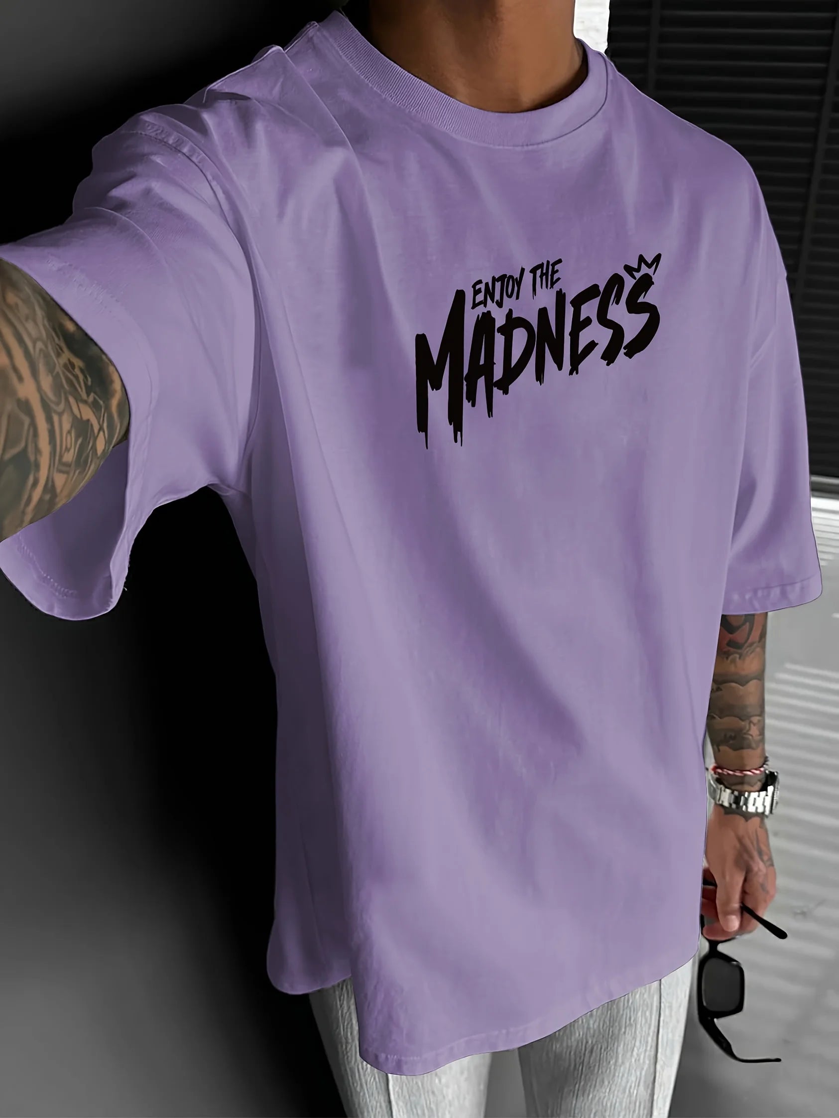 Person wearing a purple t-shirt with 'Enjoy the Madness' text, holding sunglasses.