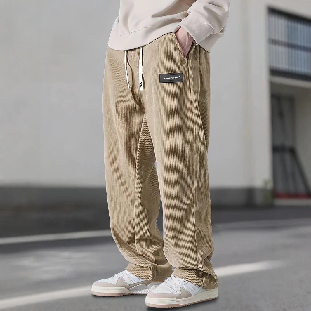 Person wearing beige corduroy pants with a brand logo, standing on a street.