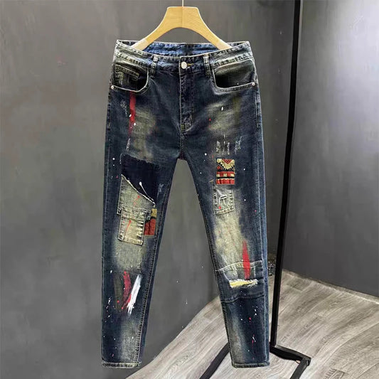 Distressed dark blue denim jeans with paint splatter, patchwork pockets, and embroidered patches, hanging on a wooden hanger against a dark gray wall.