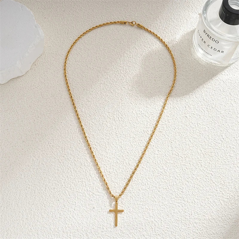 Gold necklace with a cross pendant on a textured white surface next to a Byredo perfume bottle.