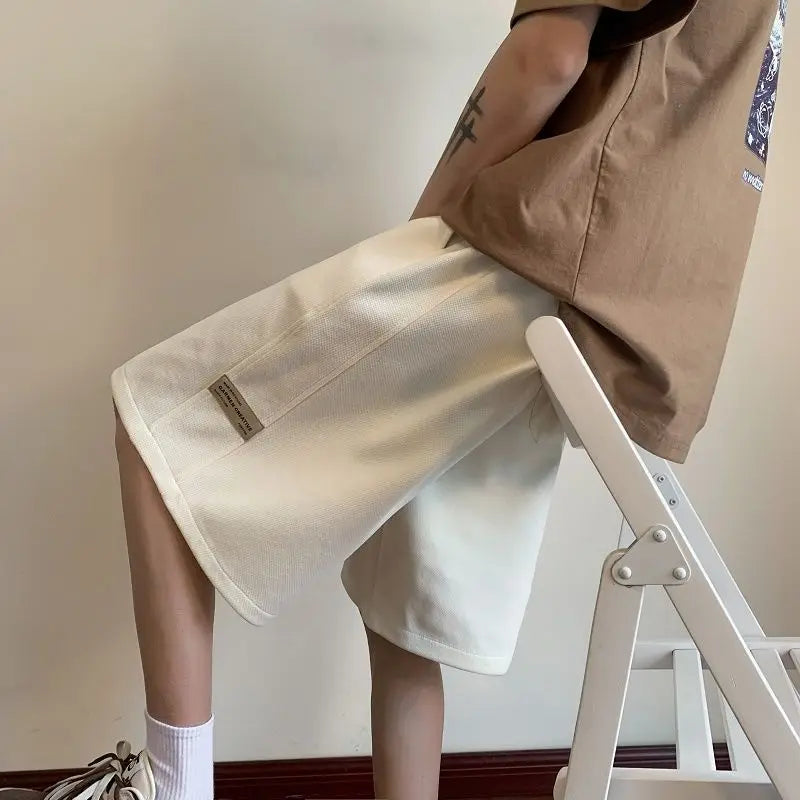 Person wearing a beige jacket and white shorts standing on a white stool against a plain wall.