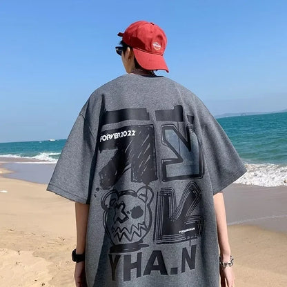 Person wearing a gray oversized t-shirt with black text and graphics on a beach.