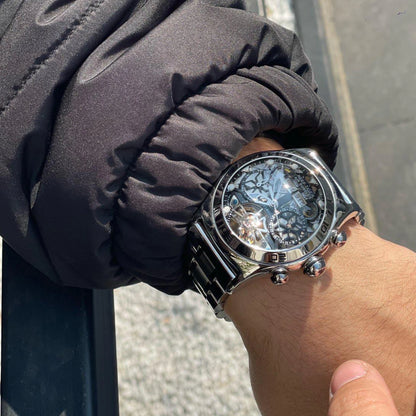 Hand wearing a silver wristwatch with a detailed face, against a blurred outdoor background.
