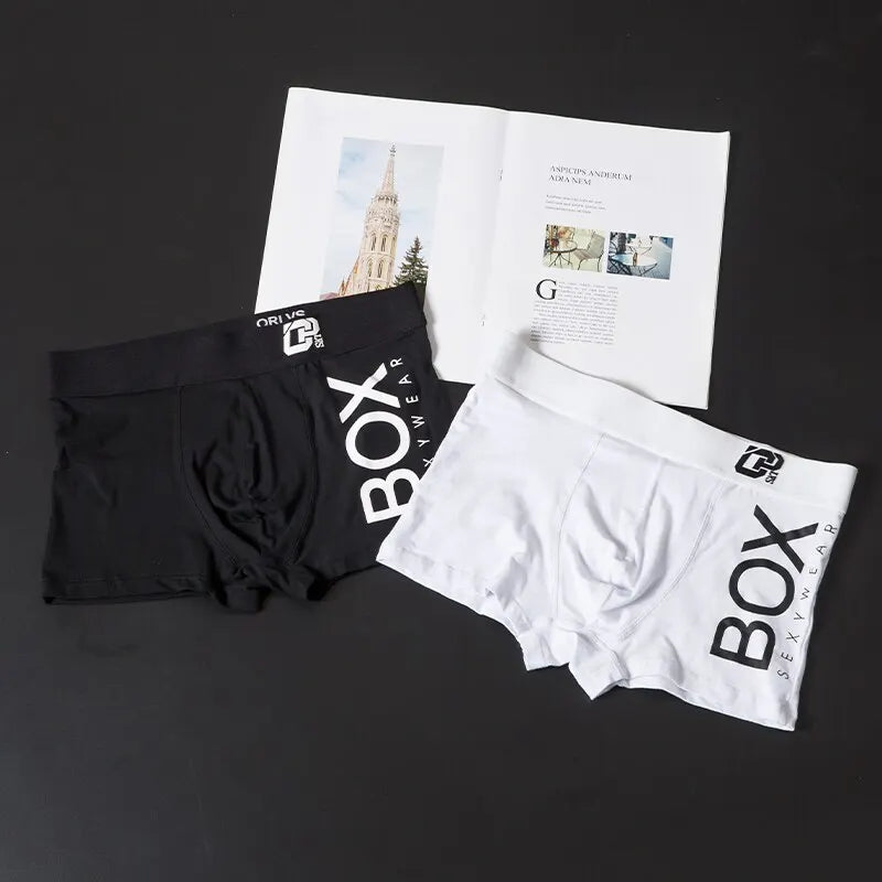 Black and white boxer briefs with 'BOX' branding on a dark surface with a newspaper.